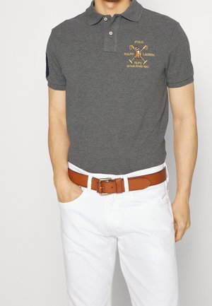 Mężczyzna ubrany w szarą koszulkę Polo Ralph Lauren z wyszywanym logo, białe spodnie i brązowy skórzany pasek, stojący z jedną ręką w kieszeni.