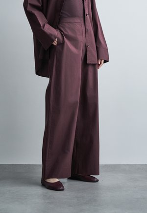 Personne portant un pantalon large bordeaux ample, une chemise oversize boutonnée assortie, et des chaussures plates bordeaux, debout sur un sol gris face à un mur uni.