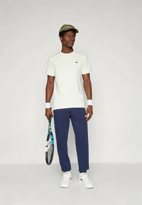 Baltā kokvilnas T-kreklā ar Lacoste logo, tumši zilos sporta biksēs, baltās kurpēs un zaļā cepurē; tur tennis raketi ar teksturētu rokturi.