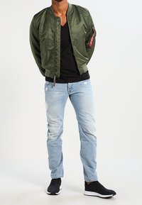 Grön satin bomberjacka med en ficka med dragkedja på sidan, svart V-ringad t-shirt, ljusblå slitna jeans och svarta sneakers.