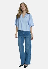 Blouse bleu clair à manches courtes et décolleté en V, associée à un jeans large de couleur bleu moyen. Le modèle porte des talons aiguilles noirs.