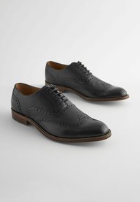 Chaussures habillées en cuir noir avec une finition texturée, perforations décoratives, bout rond et semelle marron. Présente un design à lacets.