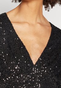 Robe noire avec un décolleté en V profond et une surface texturée ornée de sequins, créant un effet scintillant.