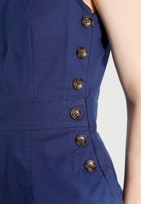 Marinblå plagg med en figursydd design, framträdande vertikala knappar längs sidan och textil med synliga sömdetaljer.