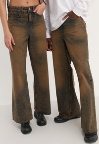 Hnědé široké džíny z denimu mají vybledlou texturu s tmavšími akcenty, opraný lem a přední kapsy, doplněné černou obuví.