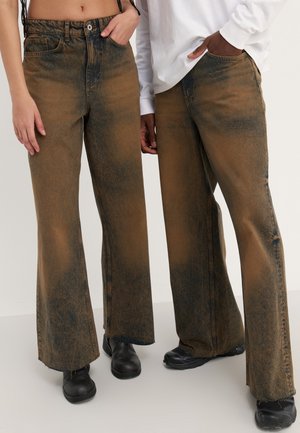 Jeans de mezclilla marrones de pierna ancha con textura deslavada y acentos más oscuros, dobladillo desgastado y bolsillos delanteros, combinados con calzado negro.