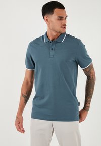 Turkizna polo majica s enotno zasnovo, belo ovratnikom in poudarki na rokavih, izdelana iz gladkega blaga. Ima dvogumbno zapiranje in kratke rokave.