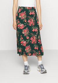 Saia midi com estampa floral em verde escuro, com flores vermelhas e rosas. Feita de tecido suave com um design fluido, combinada com sapatilhas cinzentas.