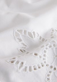 White fabric with delicate, raised floral embroidery and cut-out lace details creating a textured pattern.