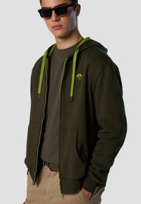 Felpa zip con cappuccio verde oliva e cordini verde lime, con tasche laterali e un piccolo logo. Indossata sopra una t-shirt scura, abbinata a pantaloni corti beige.