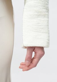 Hand relaxed downward, wearing textured white sleeve, with part of beige garment visible against plain white background.