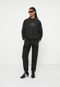 Černý sportovní outfit složený z dlouhého rukávu s kulatým límcem a logem, kalhot s elastickým pasem v odpovídající barvě a lesklých černých tenisek.