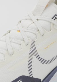 Les chaussures de trail Nike en mesh blanc présentent une surface texturée, des lacets ton sur ton et un motif géométrique rehaussé par des accents orange et une semelle bleu foncé.