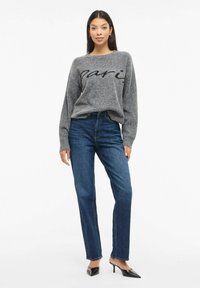 Suéter gris con letras en negro, combinado con jeans azules de tiro alto. La modelo lleva tacones negros elegantes y pendientes de aro plateados.