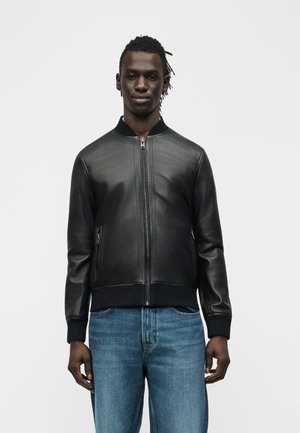 Man draagt een zwarte leren bomberjack met ritssluitingen op de zakken en een blauwe spijkerbroek, staand tegen een eenvoudige lichte achtergrond.