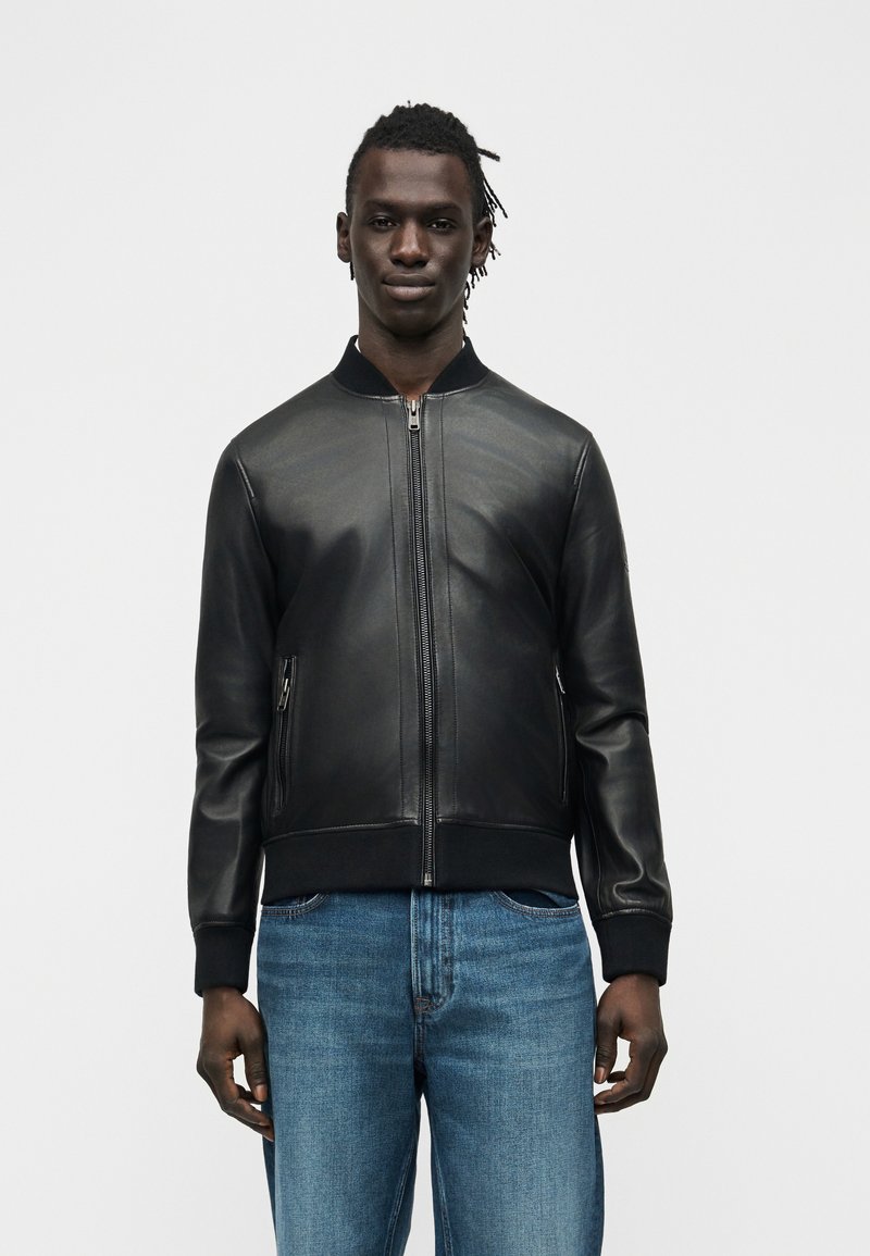 Άνδρας που φοράει μαύρο δερμάτινο μπουφάν τύπου μπόμερ με τσέπες με φερμουάρ και μπλε τζιν, στέκεται μπροστά σε απλό φωτεινό φόντο.