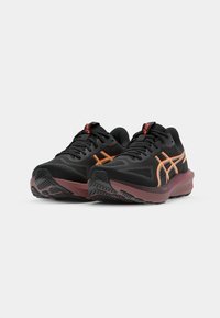 Baskets noires de sport avec des accents orange, présentant une tige en maille texturée, un col rembourré et une semelle en caoutchouc bordeaux avec un motif de traction.