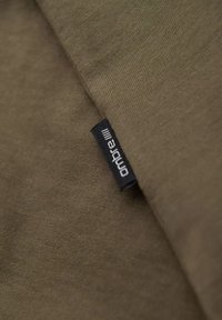 Olivově zelená látka s hladkým povrchem, na které je černá etiketa s nápisem "ombre". Na obrázku je zobrazená složená část materiálu.