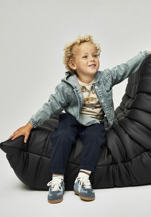 Enfant portant une veste en denim, une chemise beige à motif animal, un pantalon sombre et des baskets bleues. Assis sur une chaise en mousse noire texturée.