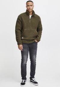 Fleece pullover in olive green, featuring a half-zip closure and front pocket. Worn with dark jeans and black sneakers.
