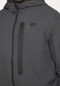 Närbild av en person som bär en strukturerad mörkgrå Nike-jacka med dragkedja fram, dragkedjeförsedd bröstficka och huva.