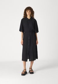 Black button-up dress with short sleeves, a tied waist, and a straight cut. Features a textured fabric and extends to mid-calf length.