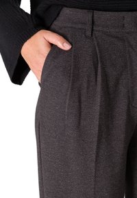 Hand mit lackierten Nägeln, in der Tasche einer dunkelgrauen, strukturierten Hose, trägt ein schwarzes, geripptes Langarmoberteil.