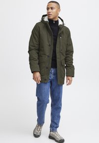Chaqueta parka con capucha de color verde oliva, con un cuello forrado de forro polar, que presenta botones y bolsillos. Combinada con jeans azules y zapatillas grises.