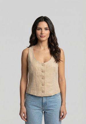 Donna con capelli lunghi e scuri che indossa un gilet beige senza maniche con bottoni e jeans azzurri, in piedi davanti a uno sfondo grigio chiaro uniforme.