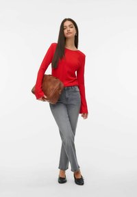 Vrouw met lang donker haar, gekleed in een rode lange mouw top, grijze jeans, zwarte schoenen, en een grote bruine leren tas in haar hand.