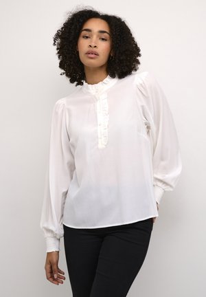 White blouse with a ruffled neckline and button placket, featuring puffed sleeves and elastic cuffs. Paired with dark fitted pants.