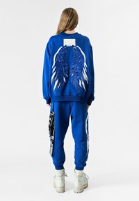 Sudadera azul con diseño de alas bordadas en la parte posterior, combinada con pantalones a juego. Presenta una etiqueta en la parte posterior y un bolsillo lateral como acento.