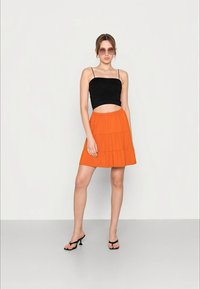 Mulher usando óculos de sol redondos, top curto preto de alças finas, saia laranja em camadas e sandálias de salto pretas, parada contra um fundo simples.