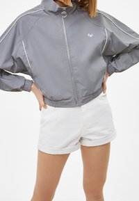 Šedá zipová bunda s bílými akcenty, s vysokým límcem a elastickými manžetami, spárovaná s bílými zkrácenými džínovými šortkami.