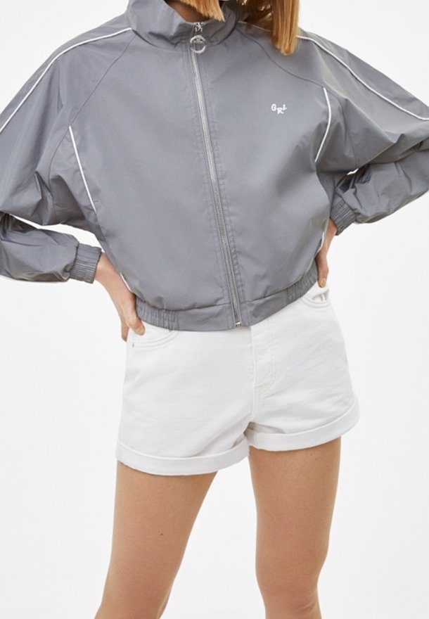 Šedá zipová bunda s bílými akcenty, s vysokým límcem a elastickými manžetami, spárovaná s bílými zkrácenými džínovými šortkami.