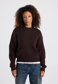 Brown knit sweater with a ribbed texture, round neckline, and dropped shoulders. Worn over a white shirt, paired with blue denim jeans.