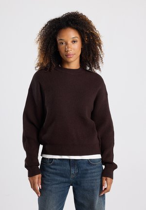 Brown gebreide trui met een geribbelde textuur, ronde halslijn en verlaagde schouders. Draag het over een wit shirt, gecombineerd met blauwe spijkerbroeken.