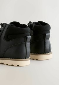 Ett par svarta läderankleboots med vadderade kragar, beige gummisålar och draflikar bakpå, på en ljus bakgrund.