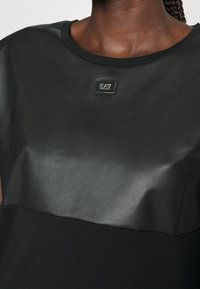 Lähedalt vaade mustale topile, millel on mati ja läikiva tekstuuriga osad ning väike ristküliku kujuline EA7 Emporio Armani logo plaastrike keskne lõikepiiril.