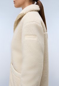 Jacke de fleece de cor creme com um acabamento texturizado, apresentando um colarinho alto e um emblema na manga. Inclui um bolso frontal.