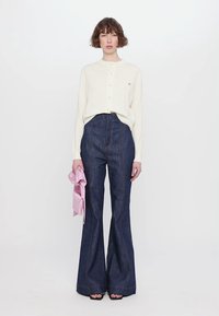 Gräddfärgad cardigan med knappar, högmidjade mörka denimbyxor med utsvängd nederkant och en rosa clutchväska i handen. Släta texturer och avslappnad design.