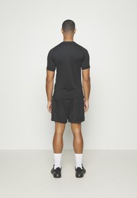Sort kortærmet atletisk top med en glat overflade, parret med sorte shorts. Modellen står med ryggen til, iført hvide sokker og sorte sko.