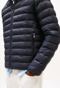 Personne portant une veste matelassée bleu marine par-dessus une chemise rayée, les mains dans les poches d'un pantalon crème, vue de la poitrine aux hanches.