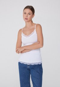 Biała koszulka na ramiączkach z koronkowym wykończeniem przy dekolcie i u dołu, z cienkimi ramiączkami. Modelka łączy ją z niebieskimi spodniami o wysokim stanie.
