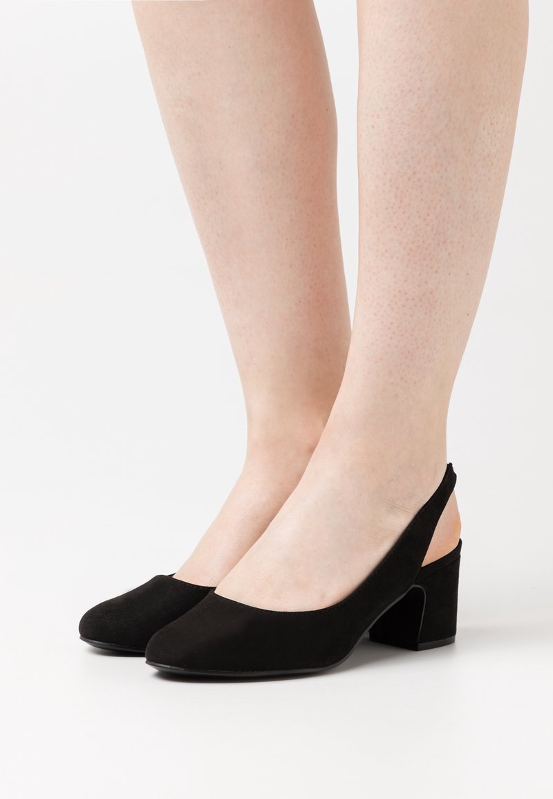 Escarpins à brides arrière en daim noir avec talons carrés portés pieds nus sur fond blanc.