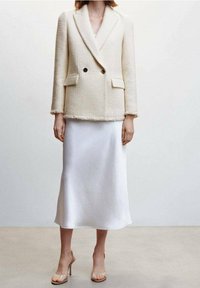 Cremfarbener, texturierter, doppelt geknöpfter Blazer mit zwei Taschen und fransigem Saum; kombiniert mit einem glatten, weißen Satin-Midirock und transparenten High Heels.