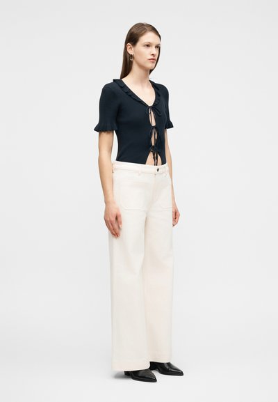 Γυναίκα που φοράει μαύρο κοντομάνικο τοπ με τρεις δέστρες μπροστά και εκρού φαρδιά παντελόνια, στέκεται μπροστά σε ένα απλό φωτεινό φόντο.