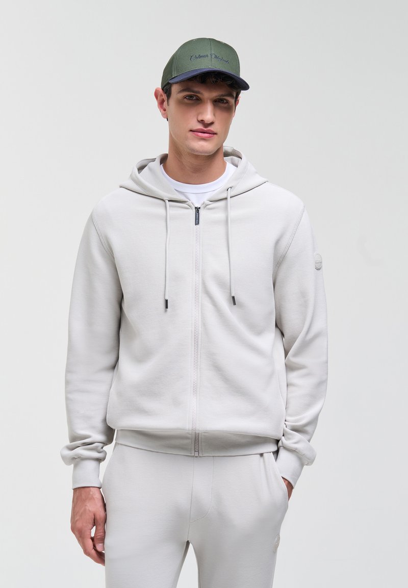 Jeune homme portant un sweat à capuche gris clair zippé et un pantalon assorti, avec une casquette de baseball vert olive, debout devant un fond uni.