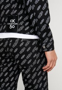 Svart denimjacka med vita "CK50"-logotyper i ett upprepat mönster, med en etikett på baksidan och en knappdetalj på ärmen.