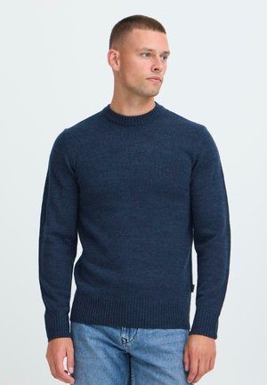 Hombre con cabello corto, vestido con un suéter de punto azul marino y jeans azul claro, mirando hacia su derecha frente a un fondo blanco liso.